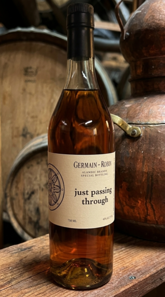 A bottle of Germain-Robin Library Single Barrel Brandy with Just Passing Through label sitting next to a small copper pot still in front of racks of barrels
